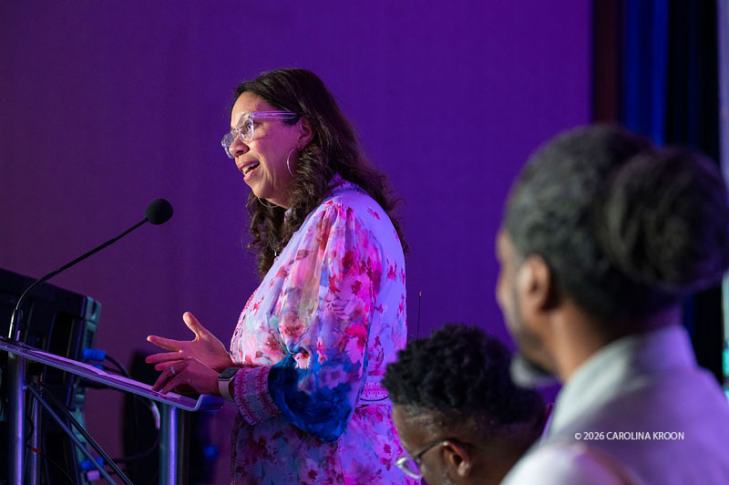 Angelique Power speaks at a podium during the GEO 2026 National Conference, smiling and gesturing with her hands. She wears a colorful pink, purple and blue dress, with fellow speakers visible in the foreground.
